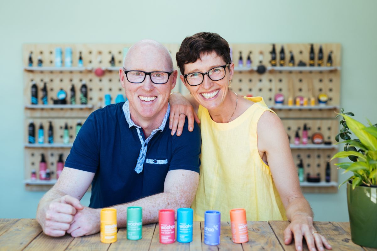 Woohoo Body co-founders Aaron and Phoebe at a table of Woohoo deodorant sticks, in front of a wall of Woohoo products